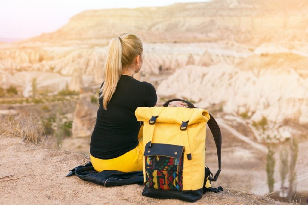 Back view blond hair woman with yellow authentic backpack looking on mountains valley. Travel adventures in desert. Goreme, Cappadocia, Turkey.  Copy space background, empty place for text.