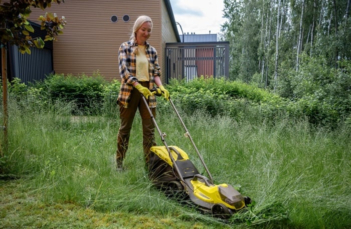 Woman,Using,Electric,Lawn,Mower,In,Overgrown,Garden,Near,Wooden